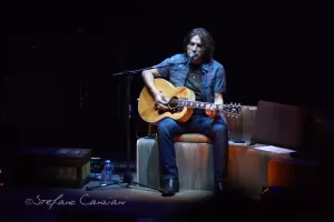 Luciano Ligabue – Artista in concerto Luciano Ligabue - Musicista che suona la chitarra acustica su un palco con ambientazione intima