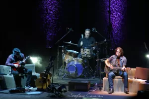 Luciano Ligabue – Esibizione dal vivo di musicisti Luciano Ligabue - Tre musicisti su un palco durante un'esibizione dal vivo con chitarra, batteria e effetti luminosi