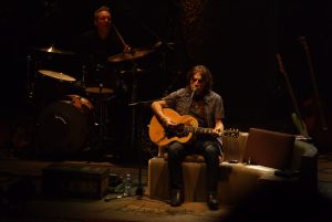 Luciano Ligabue – Concerto Intimo di Chitarra Acustica Luciano Ligabue - Musicista che suona la chitarra acustica durante un concerto con batterista sullo sfondo