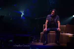 Luciano Ligabue – Concerto del musicista Luciano Ligabue - Un musicista seduto su un divano durante un concerto, con luci soffuse sullo sfondo