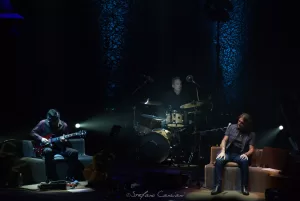 Luciano Ligabue – Trio Musicale in Performance Luciano Ligabue - Un trio musicale che suona sul palco, con chitarra, batteria e voce in un ambiente suggestivo