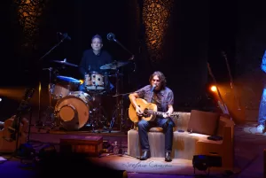 Luciano Ligabue – Concerto di Chitarra Acustica Luciano Ligabue - Artista che suona la chitarra acustica durante un concerto con batterista sullo sfondo
