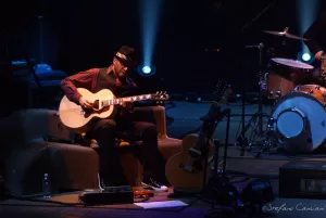 Luciano Ligabue – Chitarra sul Palco Luciano Ligabue - Musicista che suona la chitarra su un palco con luci soffuse e strumenti musicali
