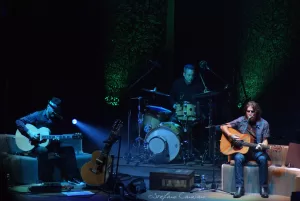 Luciano Ligabue – Esibizione musicale dal vivo Luciano Ligabue - Tre musicisti in un'esibizione dal vivo, con chitarre e batteria in un ambiente luminoso