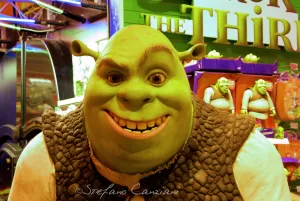 Statua di Shrek sorridente in un parco a tema, circondata da giocattoli di Shrek