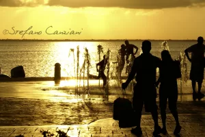 Gioco d’acqua al tramonto Persone che giocano in una fontana al tramonto, con silhouette di adulti e bambini