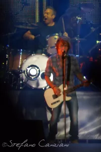 Luciano Ligabue – Concerto di rock dal vivo Luciano Ligabue - Un cantante rock con chitarra su un palco fumoso mentre un batterista suona dietro di lui