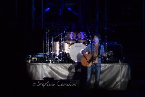 Luciano Ligabue – Concerto dal Vivo Luciano Ligabue - Un musicista su un palcoscenico con chitarra accompagnato da una band e batteria sullo sfondo