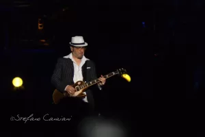 Luciano Ligabue – Musicista in azione Luciano Ligabue - Un musicista con cappello bianco suona la chitarra elettrica sul palco con luci soffuse