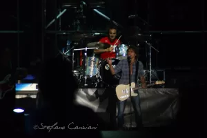 Luciano Ligabue – Concerto dal vivo di band rock Luciano Ligabue - Concerto rock con chitarrista e batterista sul palco, luci soffuse e pubblico in attesa