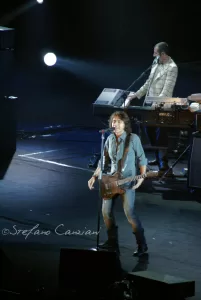 Luciano Ligabue – Concerto dal Vivo Luciano Ligabue - Un musicista sul palco con chitarra durante un concerto, con un pianista sullo sfondo