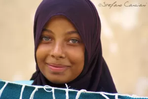 Ragazza sorridente con hijab Ragazza con hijab scuro e occhi chiari in primo piano, che sorride mentre tiene un tessuto colorato