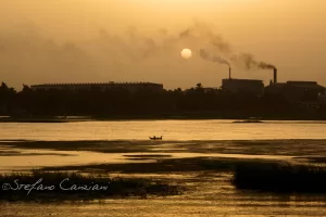 Tramonto su un fiume con una barca e fabbriche che emettono fumi