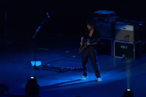Luciano Ligabue - Chitarrista in azione Luciano Ligabue - Chitarrista su un palco, illuminato da luci blu durante un concerto dal vivo