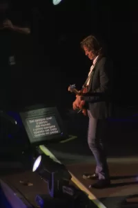 Luciano Ligabue - Artista in concerto Luciano Ligabue - Artista sul palco che suona la chitarra mentre legge il testo da un monitor.