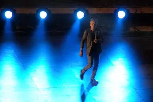 Luciano Ligabue - Uomo sul palcoscenico Luciano Ligabue - Uomo in abito elegante che cammina su un palcoscenico illuminato da faretti blu