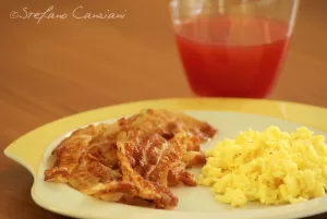 Fetta di bacon croccante accanto a uova strapazzate gialle e un bicchiere di succo di pomodoro